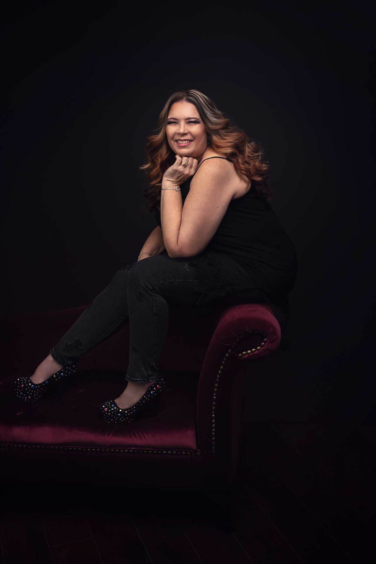 mature woman with curly hair sits on chaise in black tank top and jeans wearing fashionable jeweled shoes