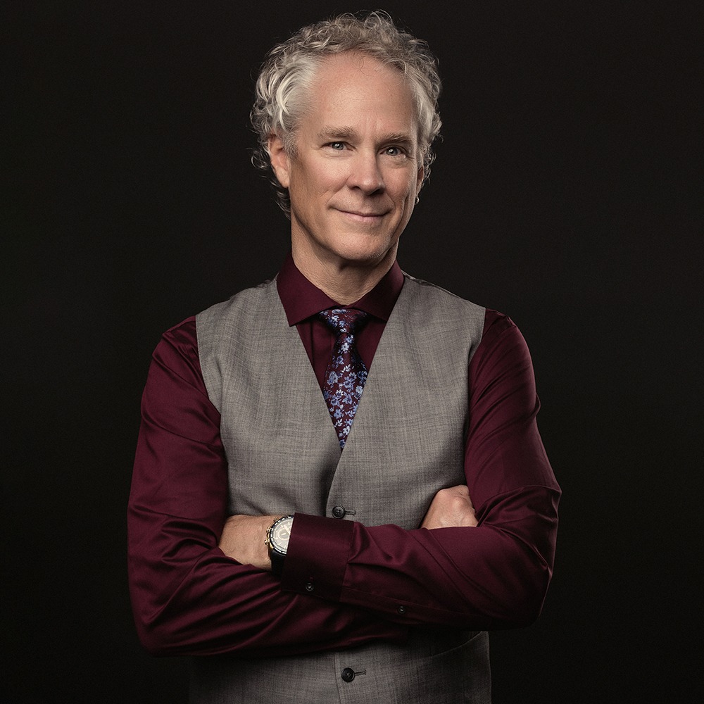 mature man with curly hair wearing maroon shirt with floral tie and grey vest