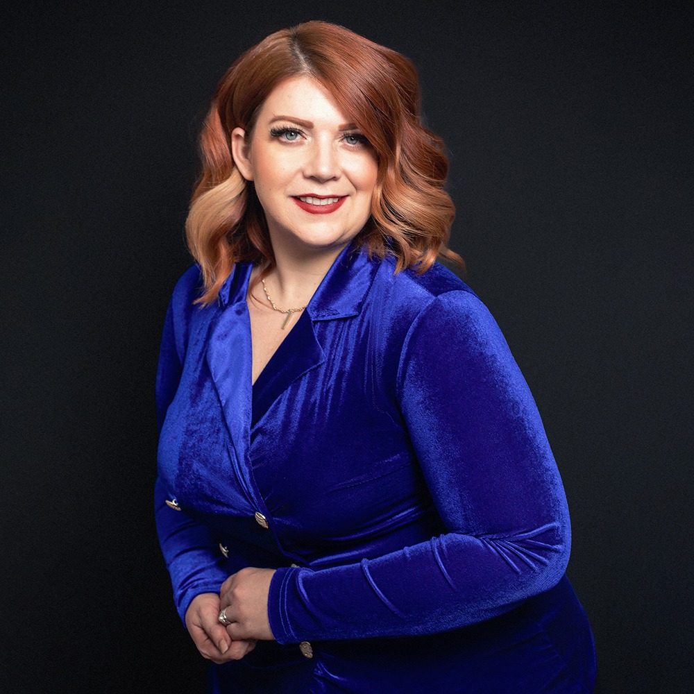 redhead wears blue velvet jacket dress in front of black background