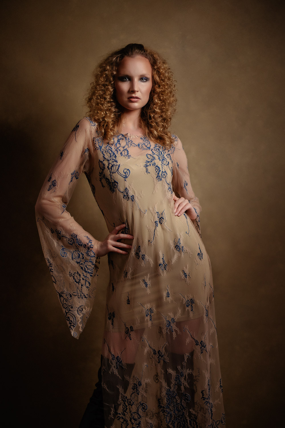 curly blonde woman striking a dramatic pose with hands on hips in sheer dress with blue embroidery
