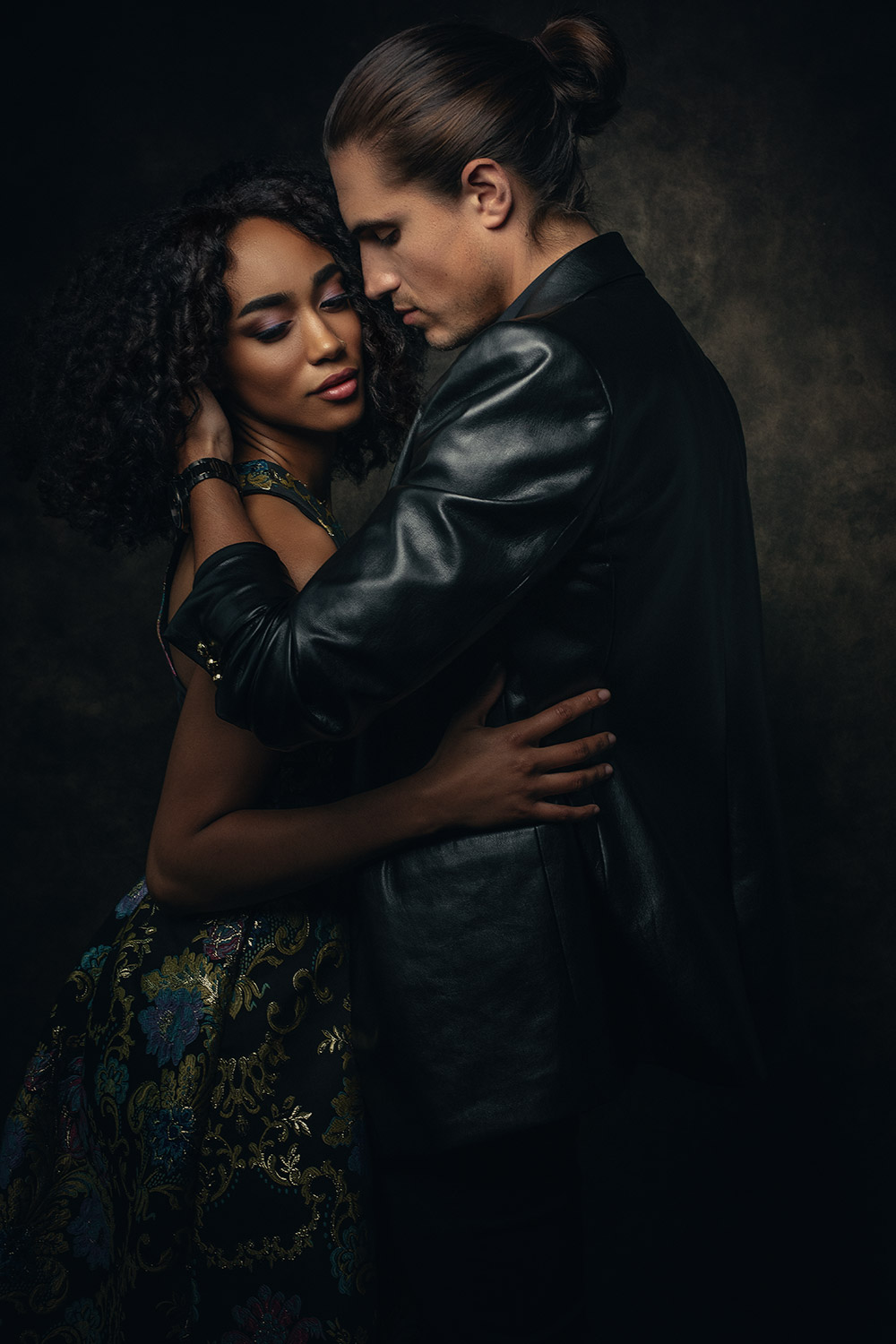 man in leather jacket embraces woman in colorful gown with gold accents in a moody lit portrait